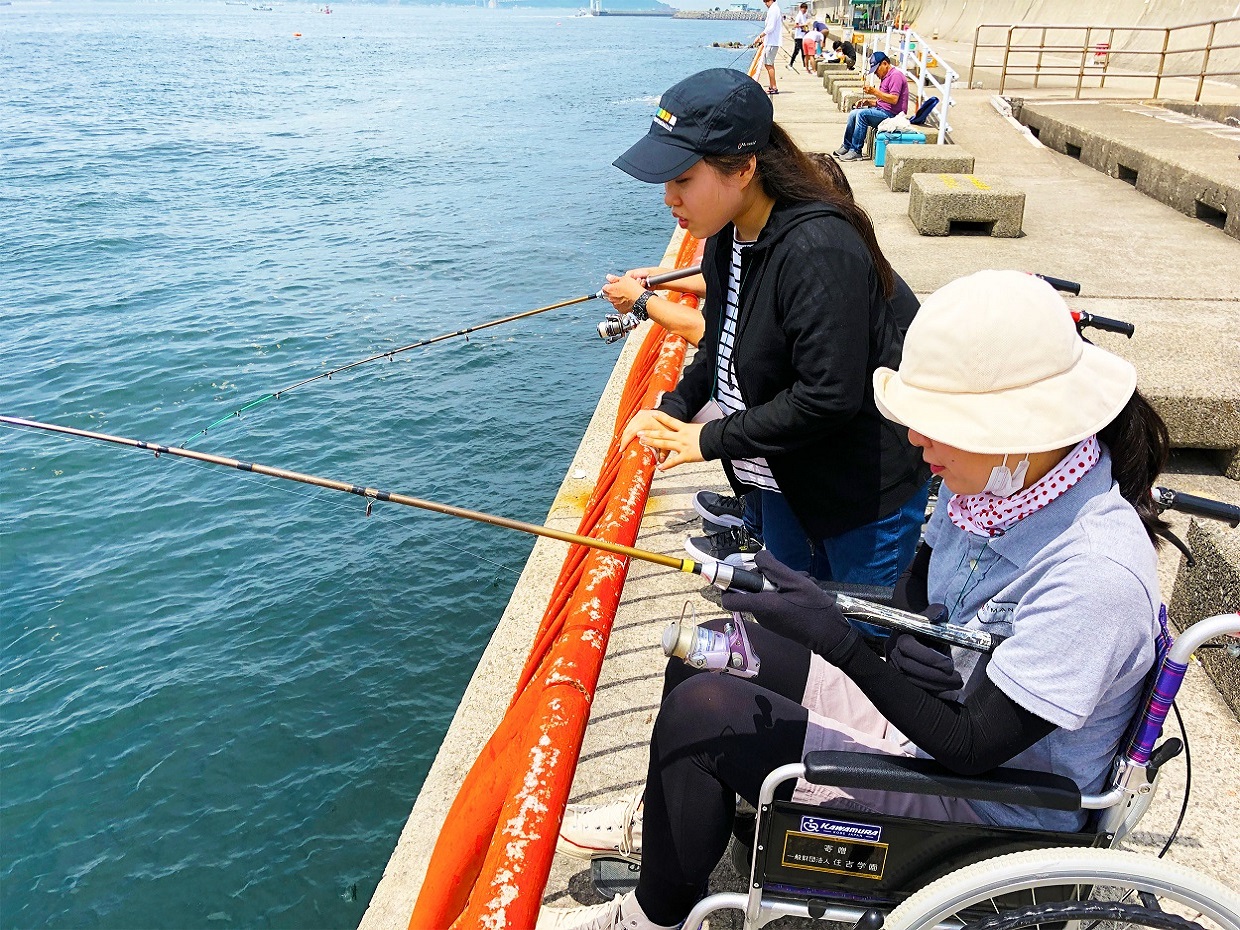 車いす体験中、竿の当たりをじっと待つ女性と、海を覗く女性
